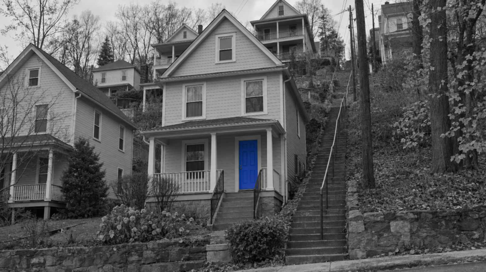Black-and-white photo of a narrow frame house on a steep Pittsburgh hillside, next to a long flight of public concrete steps. The front door is bright blue.