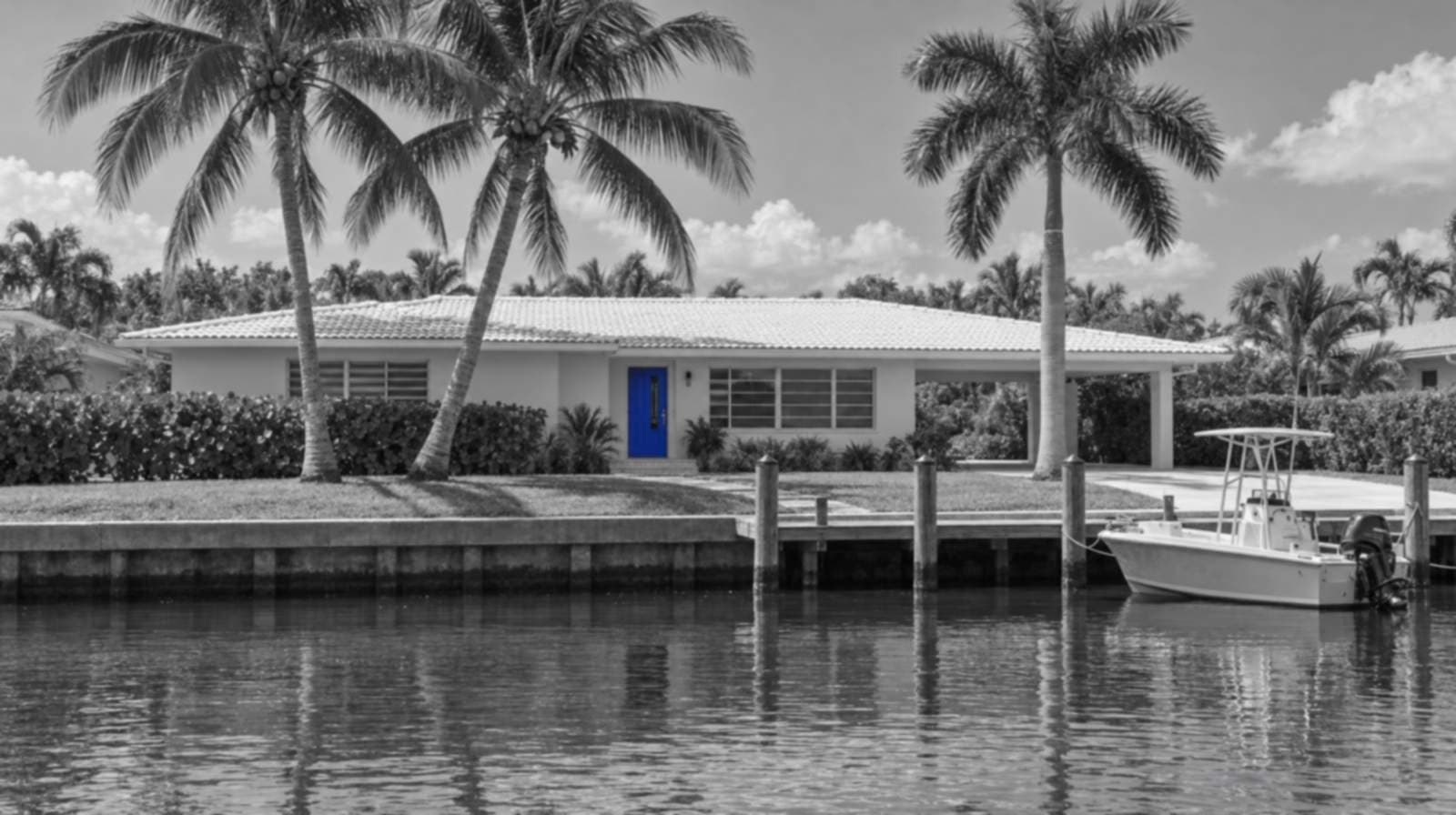 Black-and-white photo of a one-story Fort Lauderdale house on a canal, with tall palm trees, a seawall dock and a small boat. The front door is bright blue.