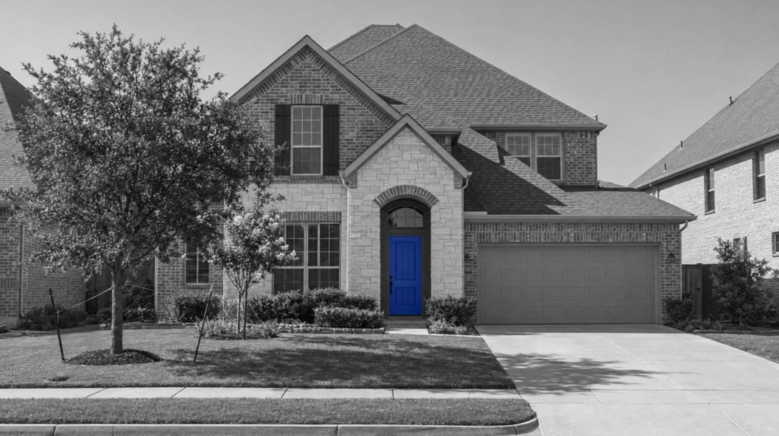 Black-and-white photo of a new two-story brick house in Plano with a stone arched entry, young staked trees and a bright blue front door.