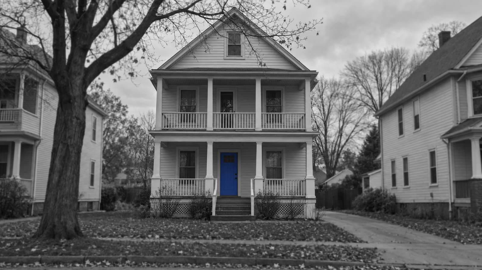 Black-and-white photo of a Cleveland two-family house with two stacked front porches, bare autumn trees and a grey sky. The front door is bright blue.