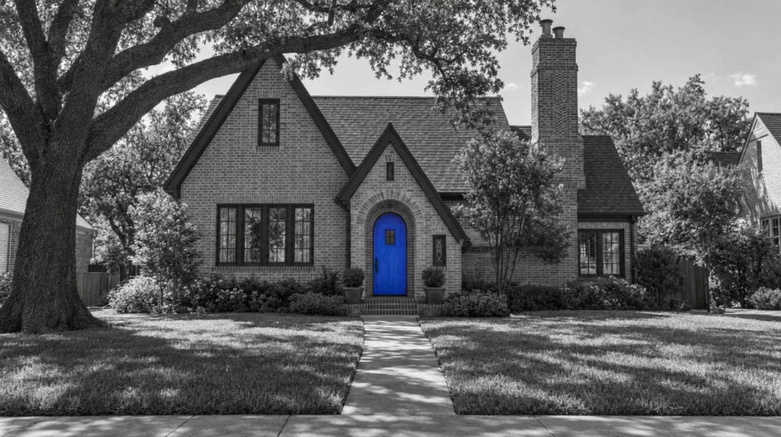 Black-and-white photo of a 1920s brick Tudor cottage in Dallas under a big live oak. Its arched front door is bright blue.