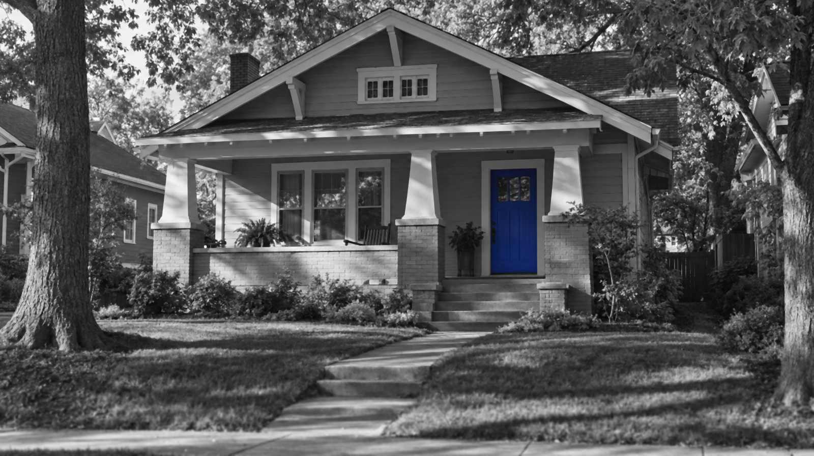 Black-and-white photo of a Nashville craftsman bungalow with a deep front porch, tapered porch columns on brick piers and tall shade trees. The front door is bright blue.