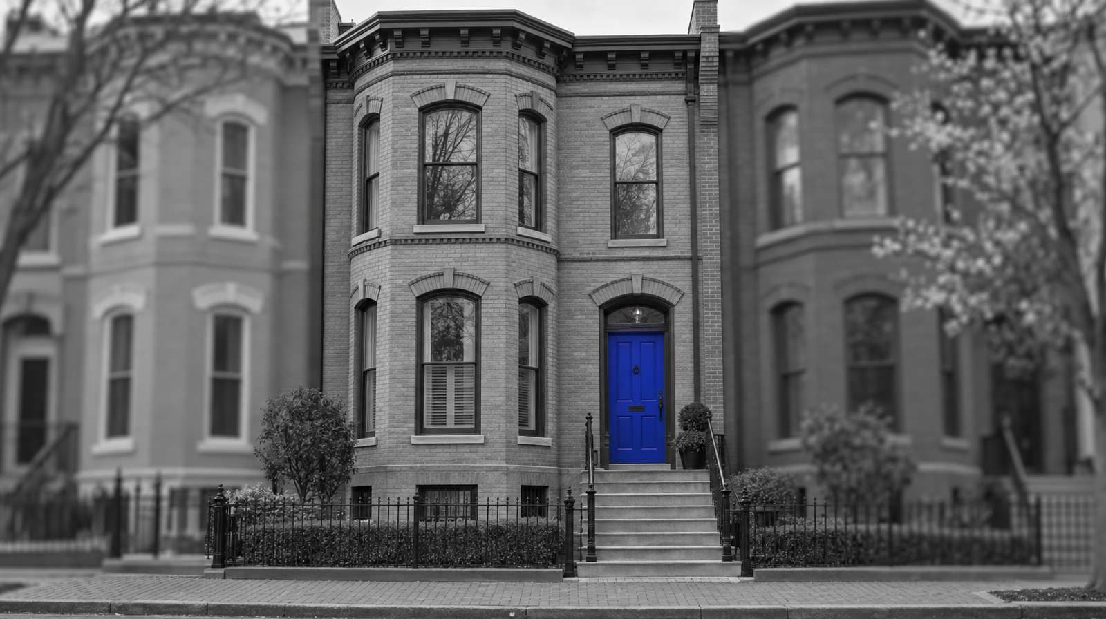 Black-and-white photo of a Victorian brick rowhouse in Washington, DC, with a bay front, stone steps and a low iron fence. The front door is bright blue.