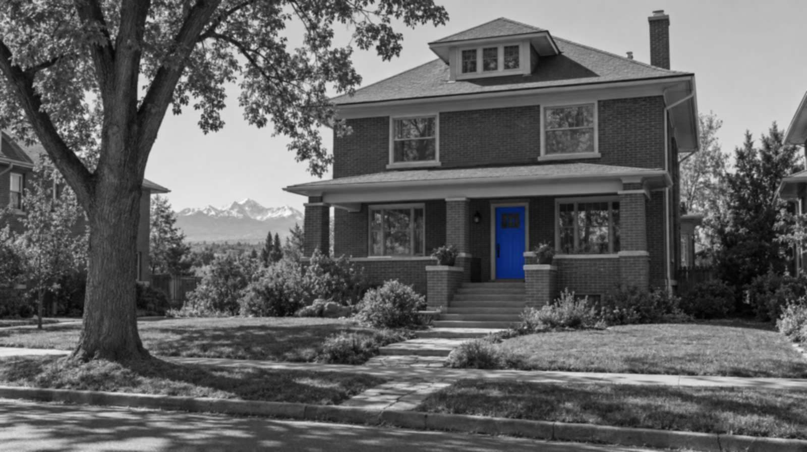 Black-and-white photo of a two-story brick Denver Square house with a wide front porch, a big shade tree and snowy mountains far behind. Only the front door is blue.