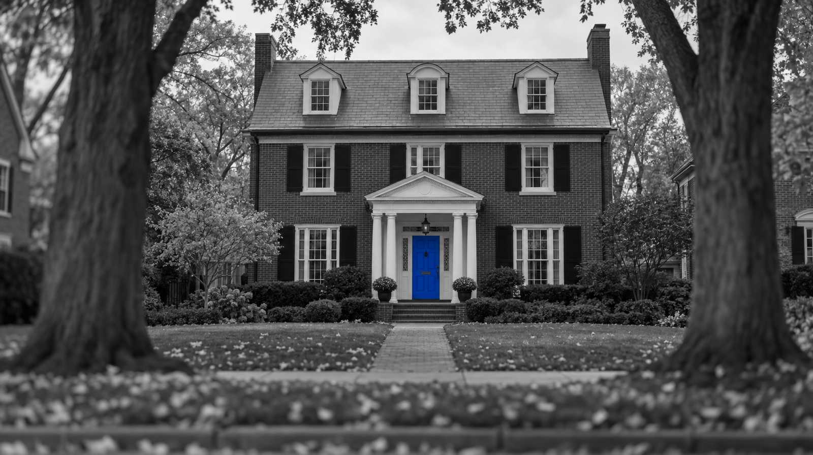 Black-and-white photo of a two-and-a-half-story brick Colonial Revival house between two large old trees, with a bright blue front door