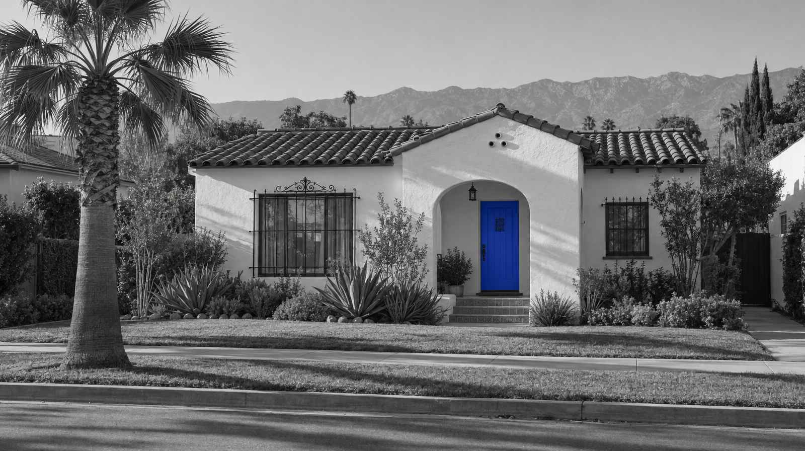 Black-and-white photo of a Spanish-style stucco bungalow with a clay tile roof, an arched porch, agave plants and a tall fan palm, with mountains in the distance. The front door is bright blue.