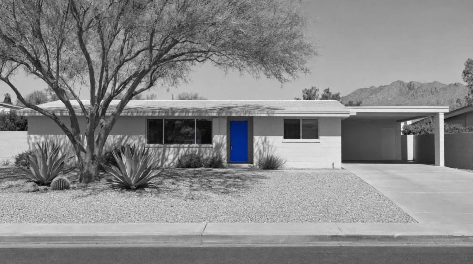 Black-and-white photo of a 1960s Phoenix ranch house built of painted block, with a carport, a gravel yard, a palo verde tree, agaves and a blue front door