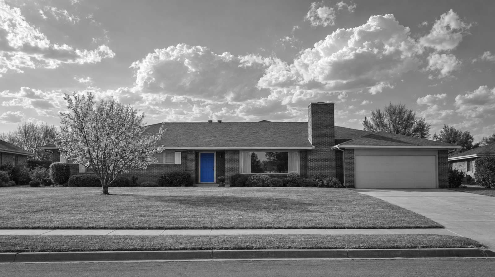 Black-and-white photo of a long brick ranch house in an Oklahoma City neighborhood, with a wide lawn, a blooming redbud tree, big clouds and a bright blue front door.