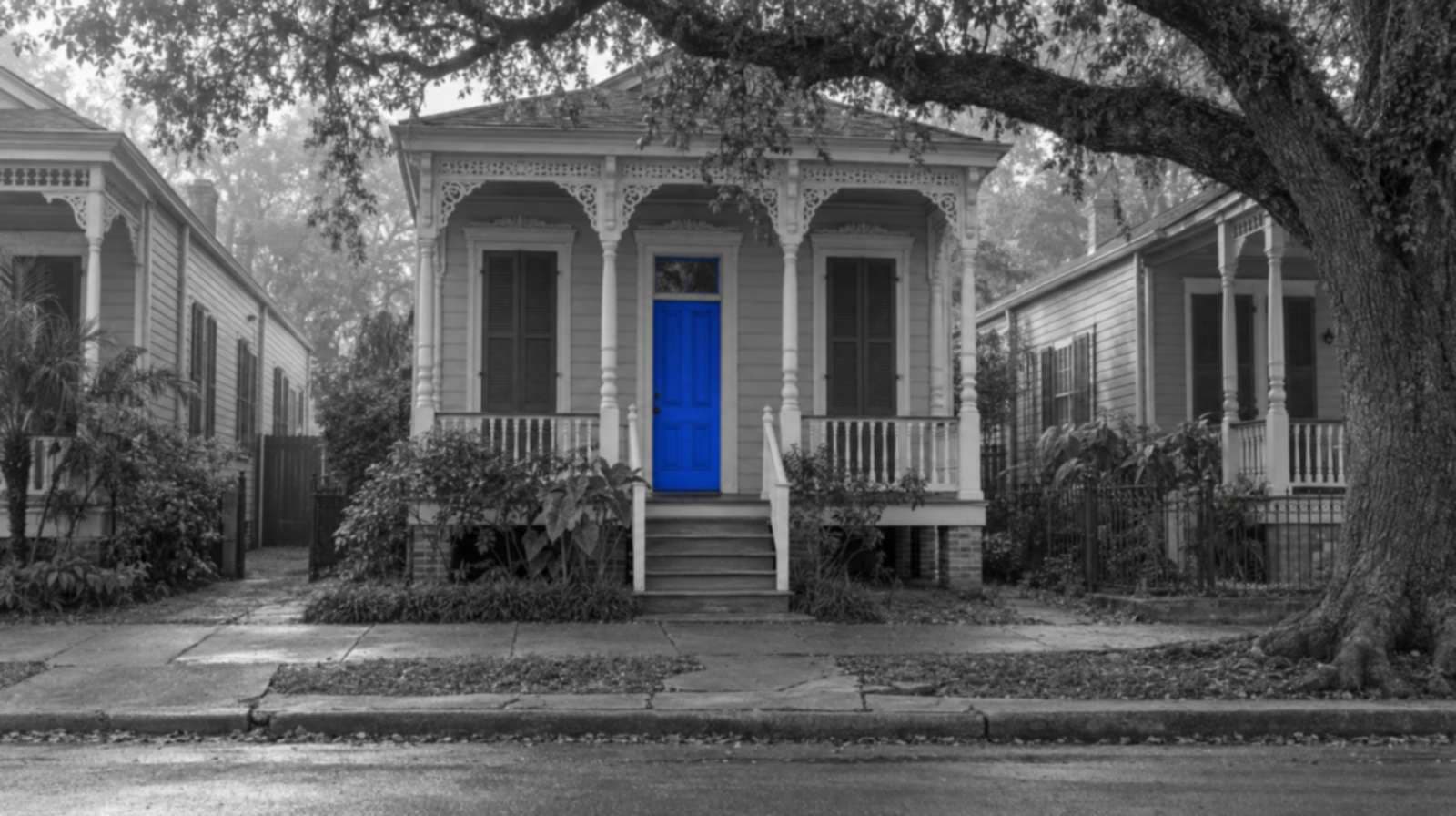 Black-and-white photo of a raised New Orleans shotgun house with gingerbread trim, shutters and a live oak over the sidewalk. Only the front door is bright blue.