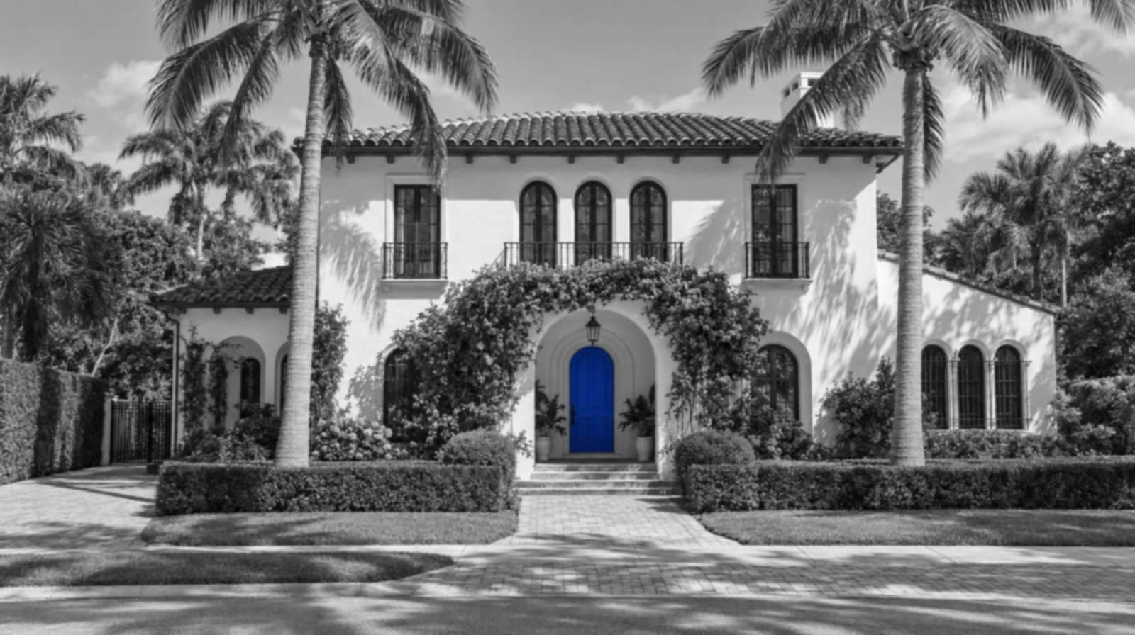 Black-and-white photo of a two-story Mediterranean Revival house in Palm Beach County, with arched windows, a barrel tile roof, bougainvillea over the entry, two coconut palms and a bright blue front door.