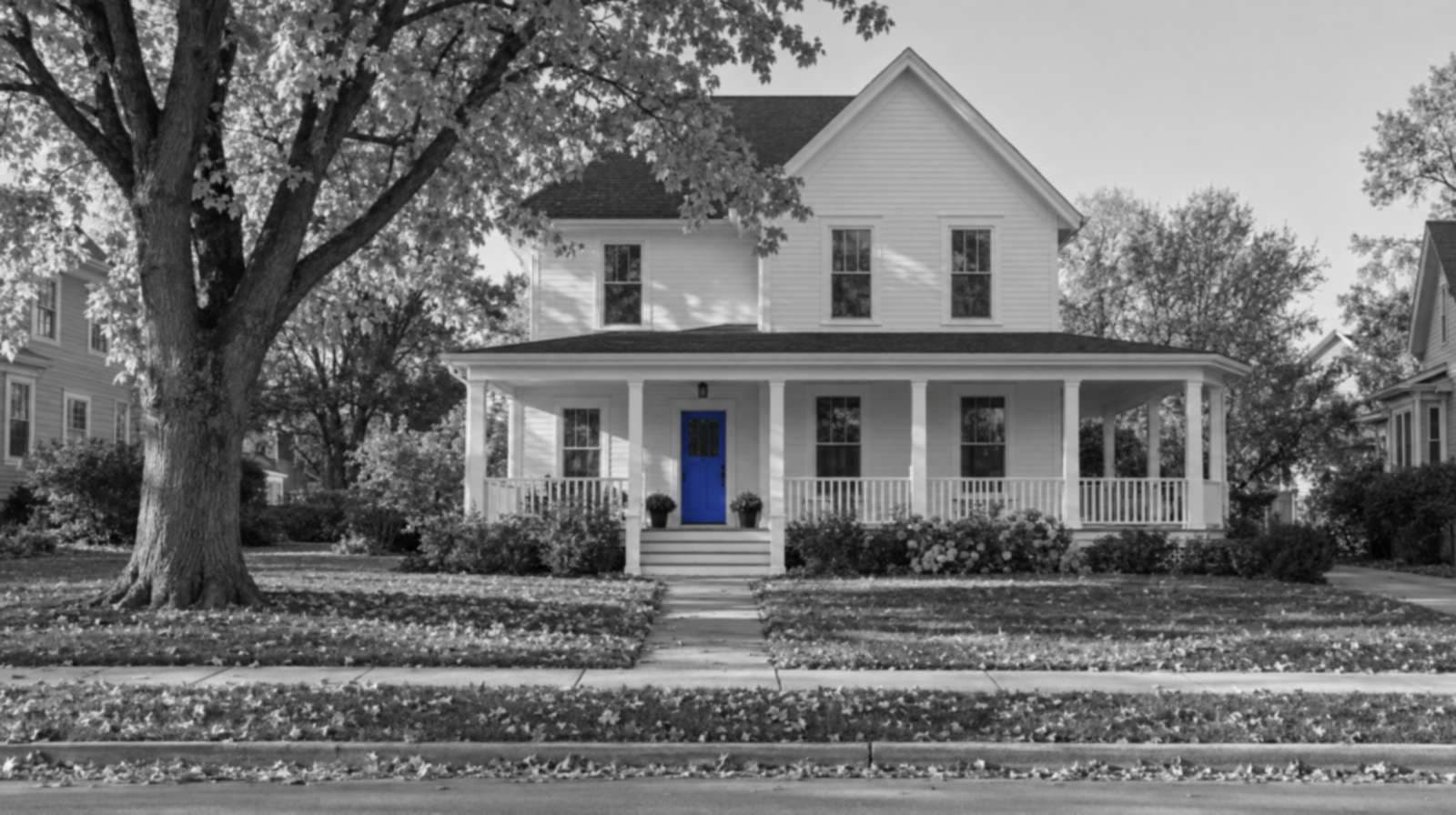 Black-and-white photo of a white two-story farmhouse-style home with a wraparound porch and a big maple tree dropping leaves on the lawn. Only the front door is bright blue.