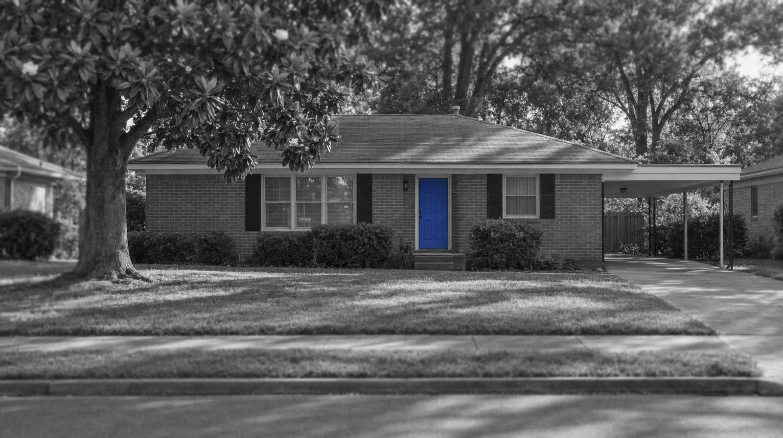 Black-and-white photo of a one-story brick ranch house in Memphis, with a side carport and a big magnolia tree in the yard. The front door is bright blue.