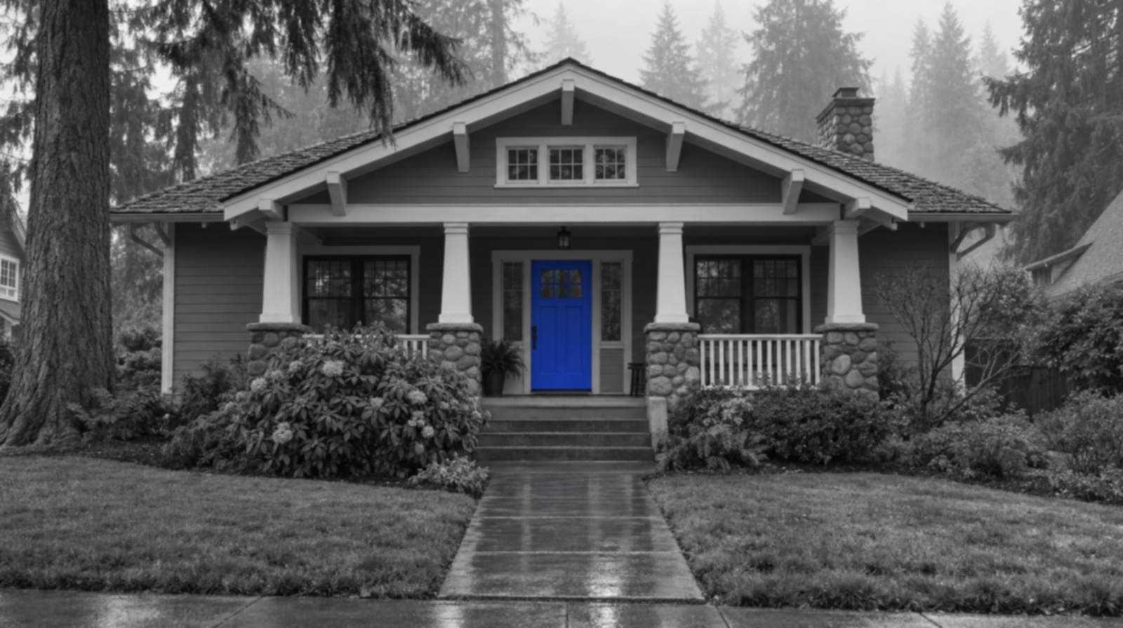 Black-and-white photo of a craftsman bungalow in Snohomish County, Washington, with river-stone porch piers, tall firs in morning mist, a wet front path and a bright blue front door.