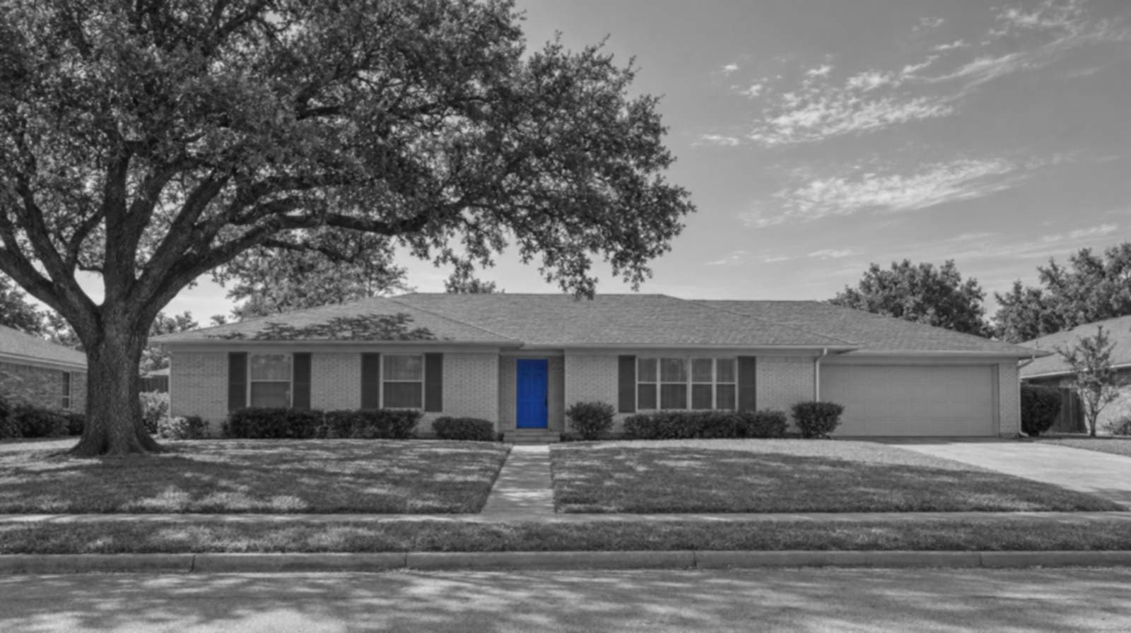 Black-and-white photo of a single-story brick ranch house with a wide lawn, a big live oak and an open Texas sky. Its front door is bright blue.