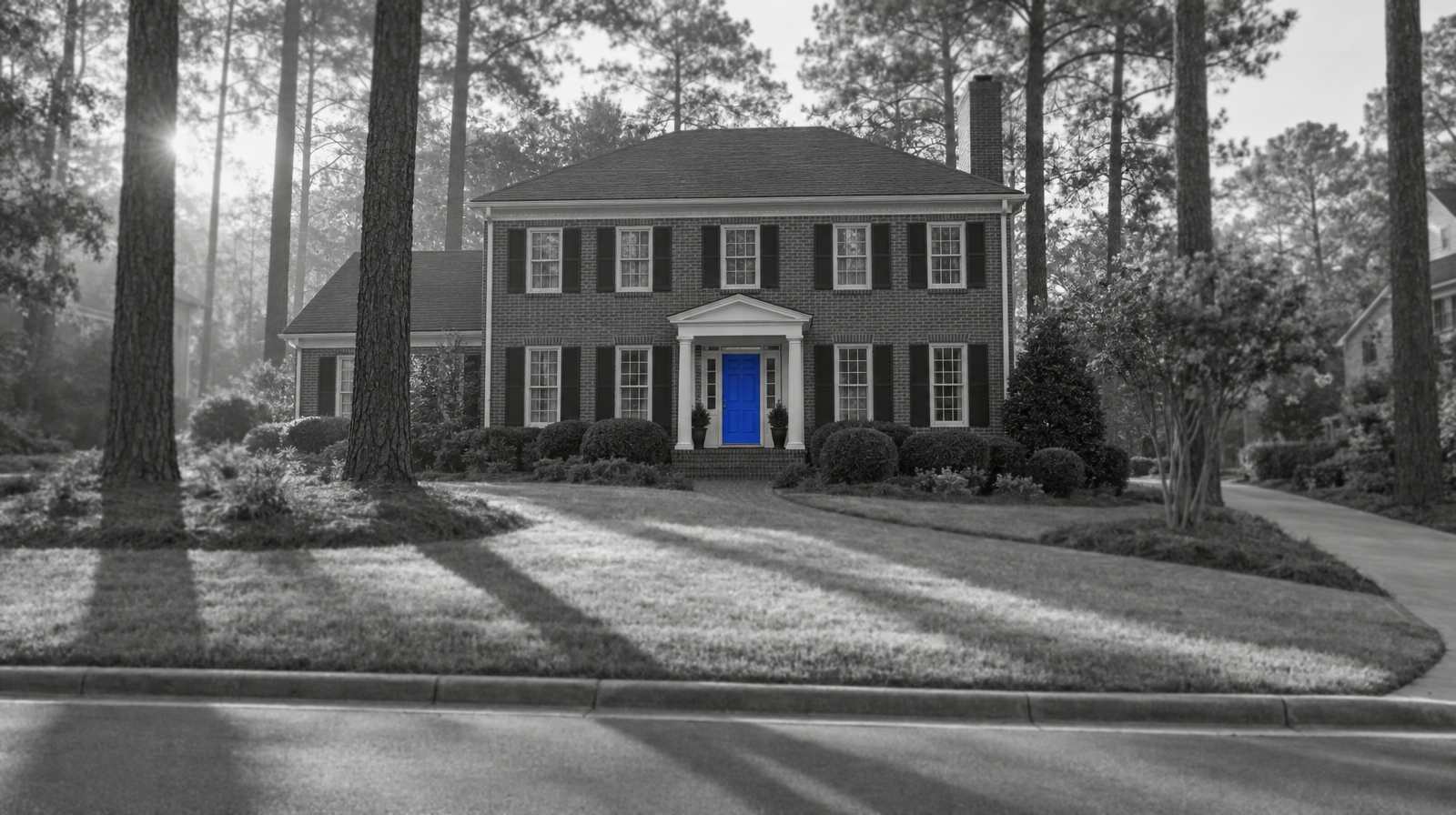 Black-and-white photo of a two-story brick colonial home under tall loblolly pines, with long morning shadows on the lawn and a bright blue front door.
