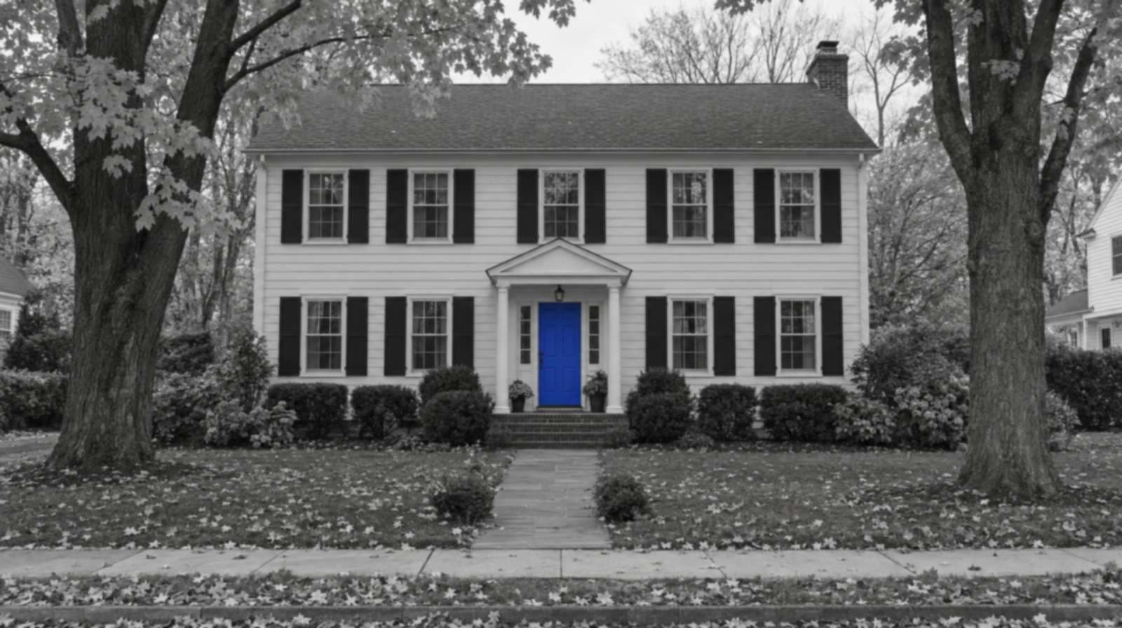Black-and-white photo of a white center-hall colonial house with black shutters, autumn maples and fallen leaves on the lawn, and a bright blue front door