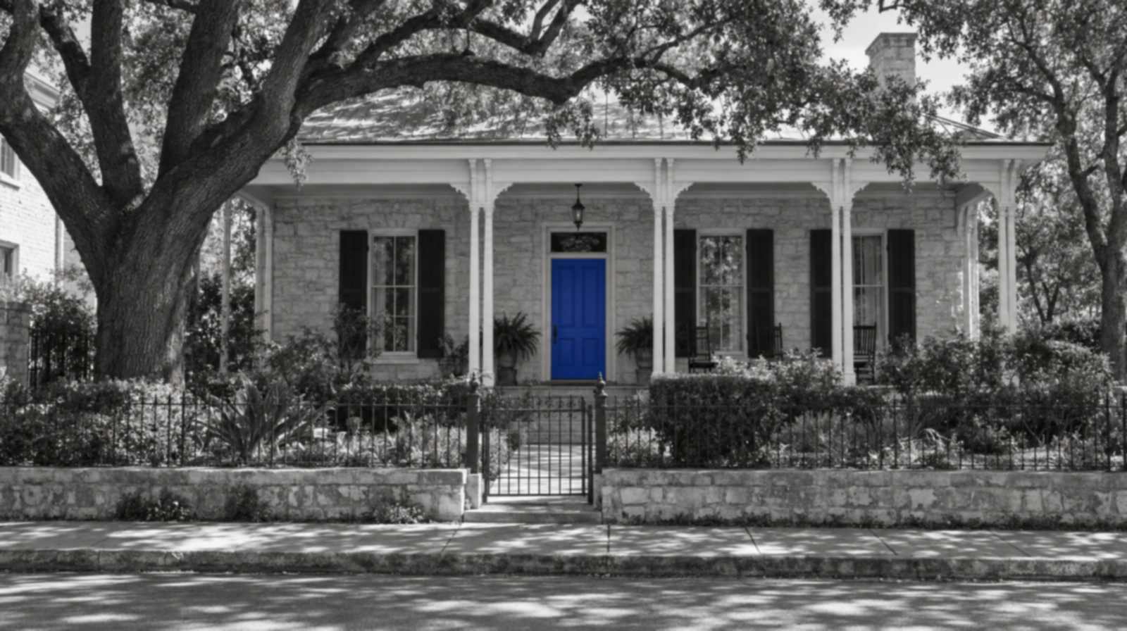 Black-and-white photo of a San Antonio style limestone house with a deep white-columned porch, a live oak and an iron fence, with one bright blue front door