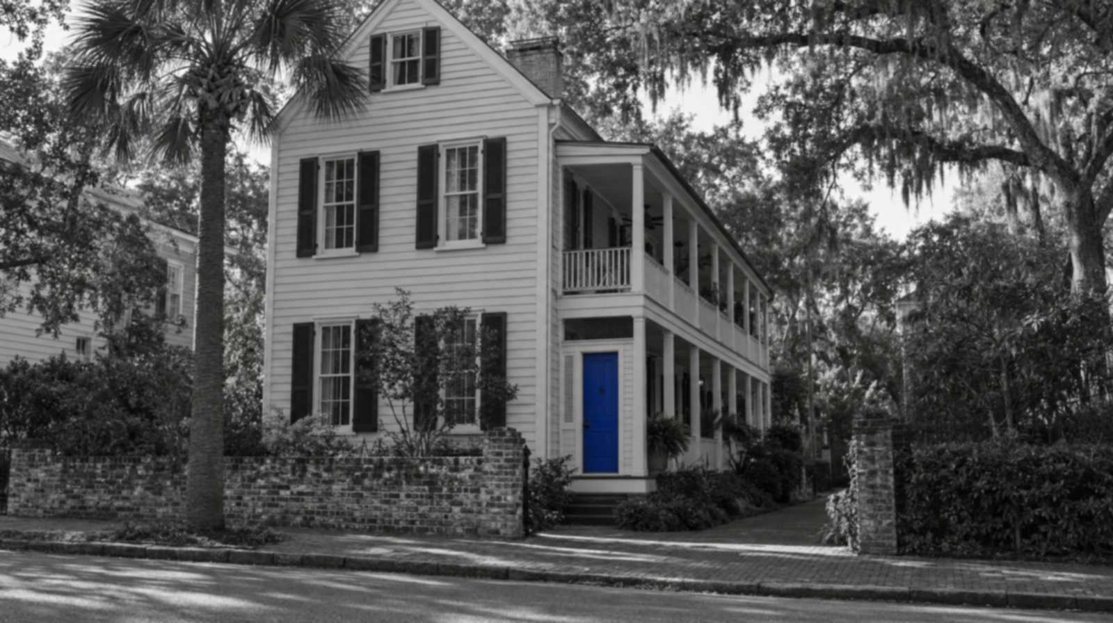 Black-and-white photo of a tall Charleston single house with a long two-story side piazza, a palmetto tree, a low brick wall and live oaks hung with Spanish moss. The piazza's street door is bright blue.