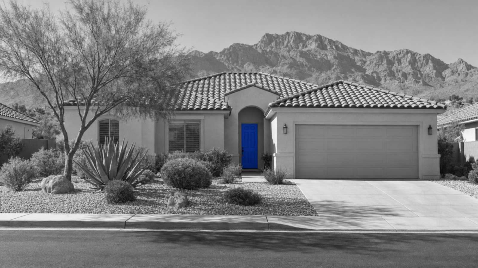 Black-and-white photo of a one-story Las Vegas stucco house with a tile roof, a gravel desert yard, an agave and rocky mountains behind it. The front door is bright blue.