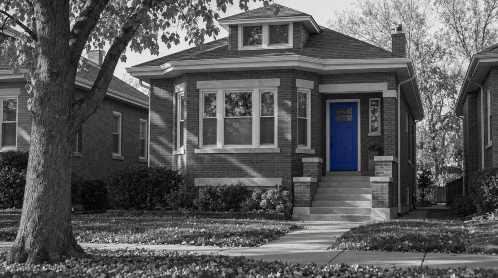 Black-and-white photo of a Chicago brick bungalow with a low hip roof, a front bay window and autumn leaves on the lawn. Only the front door is blue.