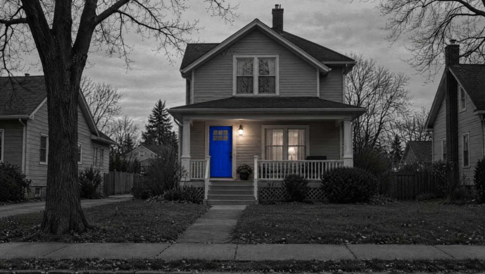 A two-story house at dusk in black and white, with its porch light on and a bright blue front door.