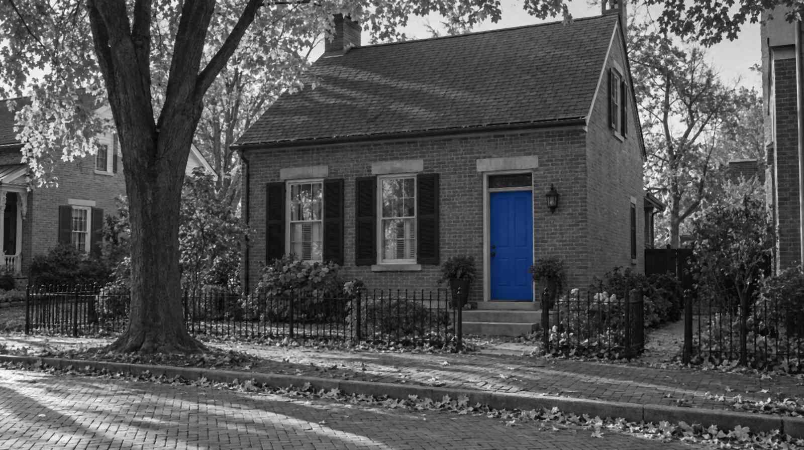 Black-and-white photo of a small red-brick cottage in Columbus's German Village, with a brick sidewalk, an iron fence and fallen autumn leaves. Only the front door is bright blue.
