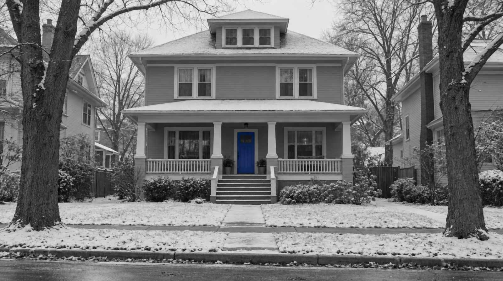 Black-and-white photo of an American foursquare house with a wide front porch, light snow on the lawn and bare elm trees. The front door is bright blue.