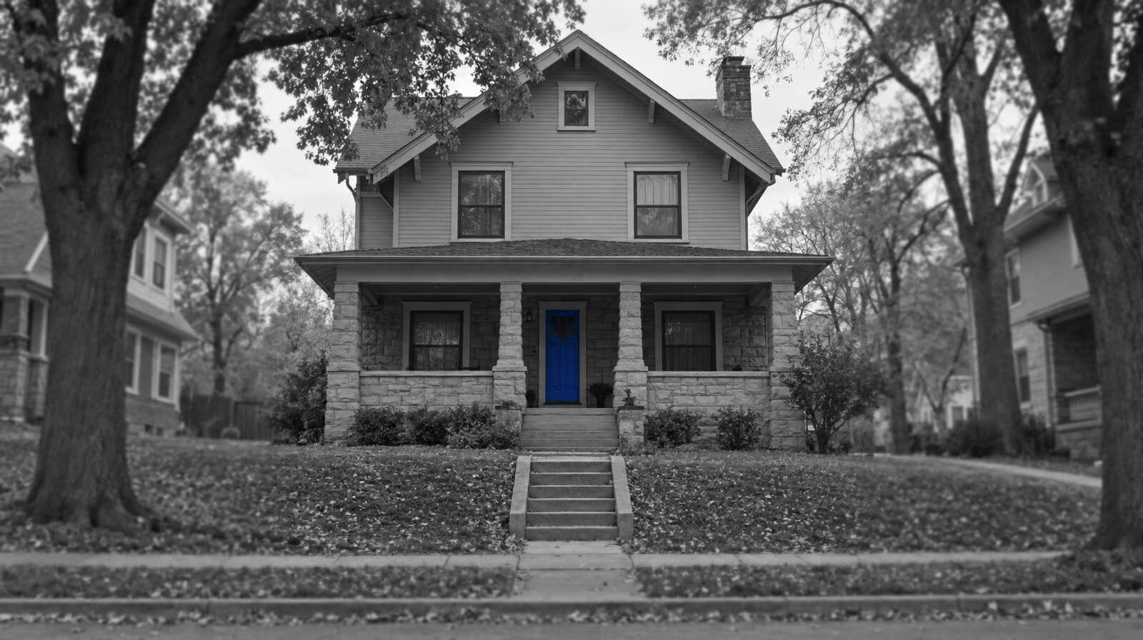 Black-and-white photo of a Kansas City shirtwaist house with a stone first floor, wood siding above and a deep front porch, on a street lined with tall trees. The front door is bright blue.