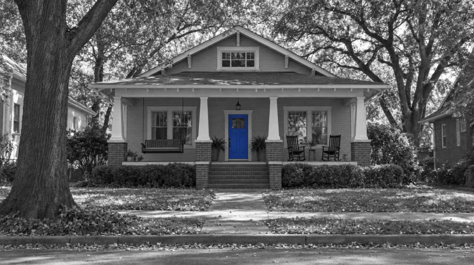 Black-and-white image of a 1920s craftsman bungalow, the kind found in Charlotte's older neighborhoods, with a wide front porch and rocking chairs under tall oak trees. The front door is bright blue.