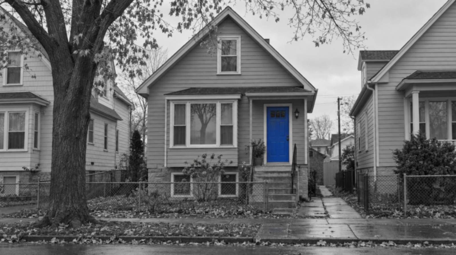 Black-and-white photo of a Milwaukee raised cottage in the local Polish flat style, with a concrete front stoop, a chain-link fence and fallen leaves under an overcast sky. The front door is bright blue.