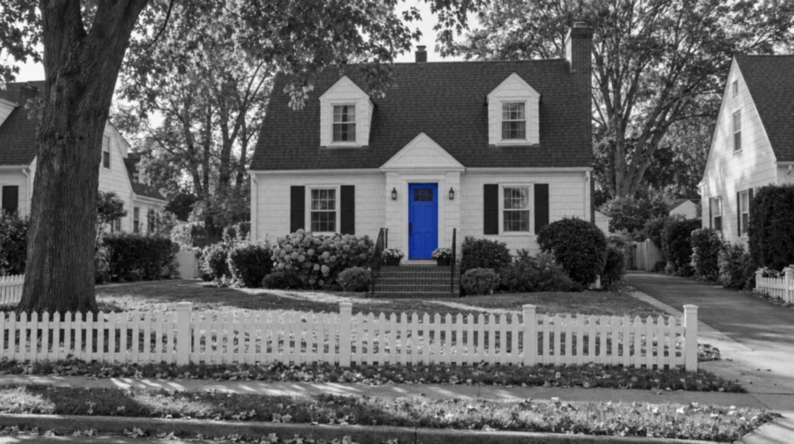 Black-and-white photo of a Long Island Cape Cod house with two dormers behind a white picket fence. Only the front door is bright blue.