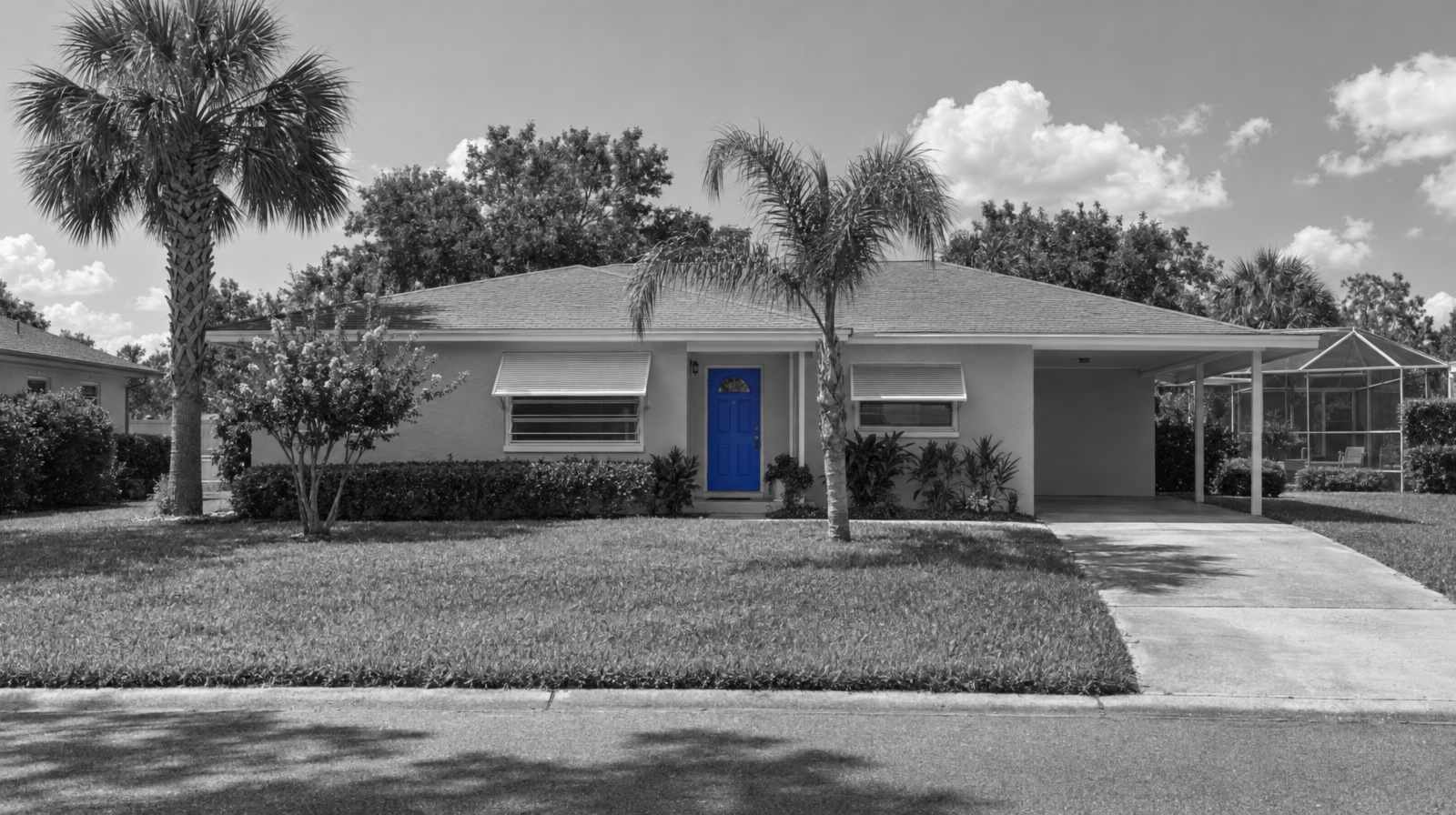 Black-and-white photo of a one-story concrete-block ranch house near Orlando, with a carport, a screened pool cage, two palm trees and a bright blue front door.