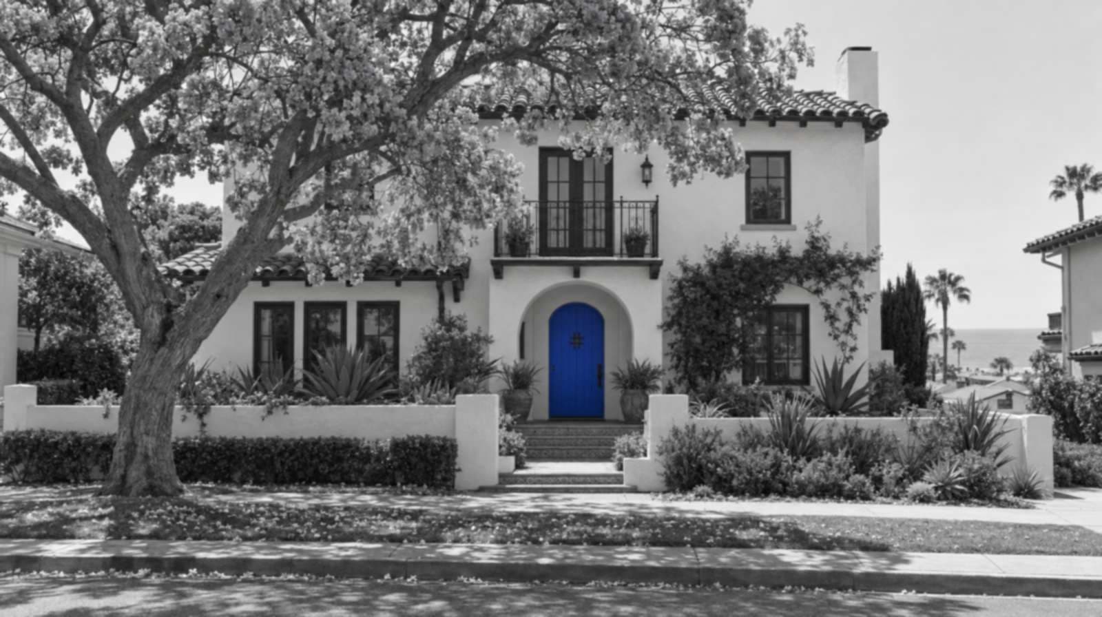 Black-and-white photo of a two-story Spanish-style stucco house with a clay tile roof, an arched porch and a small iron balcony, shaded by a blooming jacaranda tree, with palms and the ocean in the distance. The front door is bright blue.