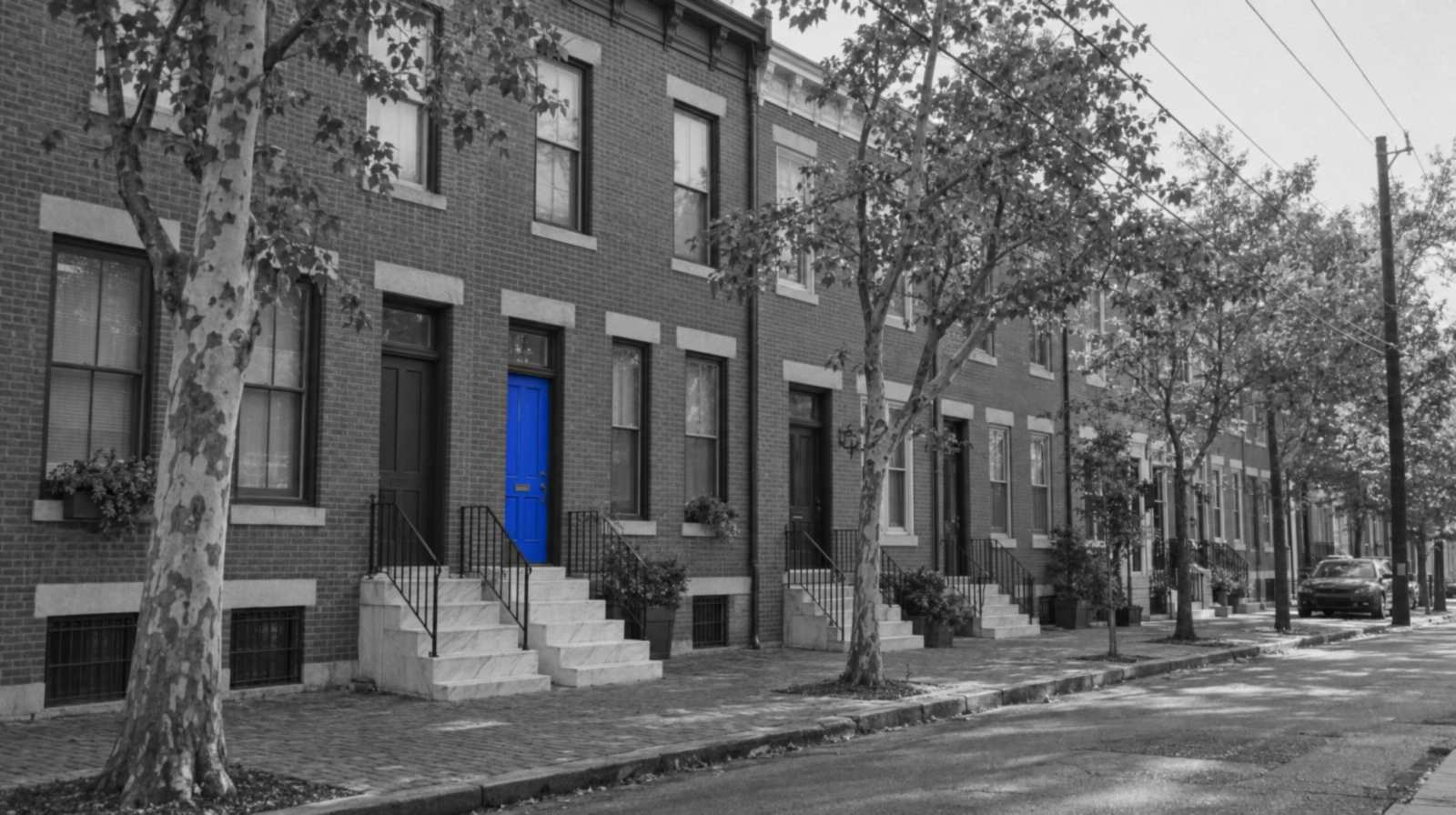 Black-and-white photo of a quiet block of Philadelphia brick rowhouses with white marble front steps and street trees. One front door is bright blue.