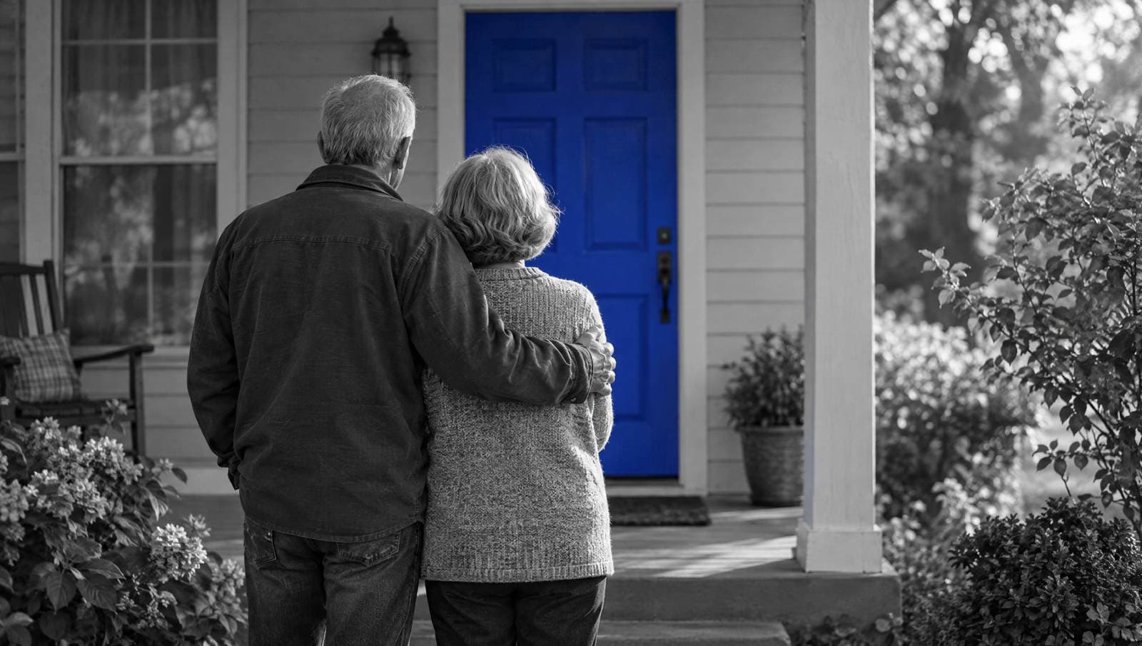 An older couple, seen from behind, stands on the porch of their home. The front door is painted blue.