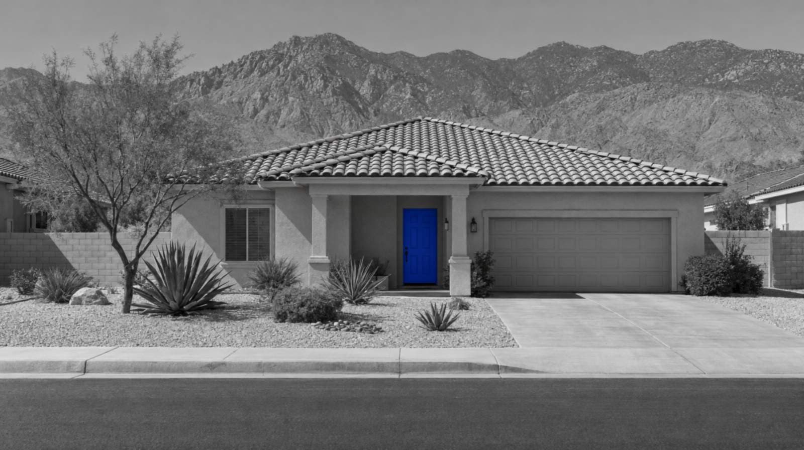 Black-and-white photo of a one-story stucco ranch house with a clay tile roof, desert plants and a blue front door, with rocky Inland Empire mountains behind it