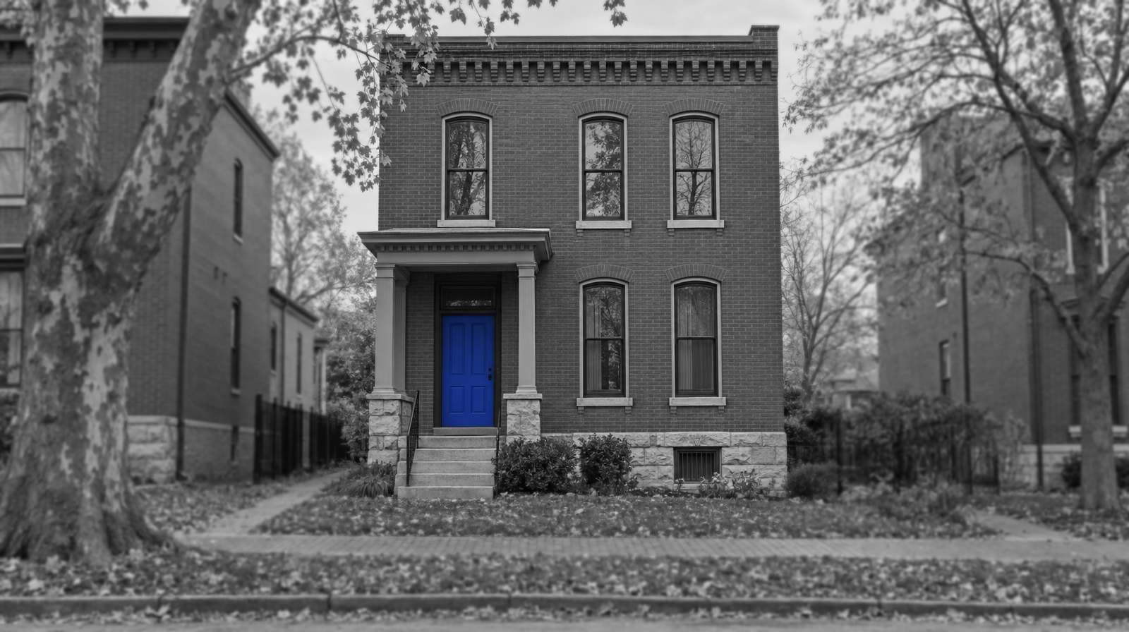 Black-and-white photo of a two-story brick St. Louis house with arched windows, a stone base and a small columned porch, between autumn trees. The front door is bright blue.